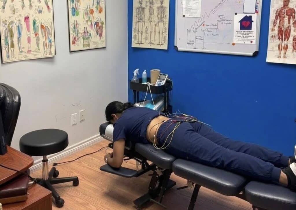 Modern physiotherapy clinic treatment room with blue walls, anatomical charts, framed credentials, and black padded treatment table.