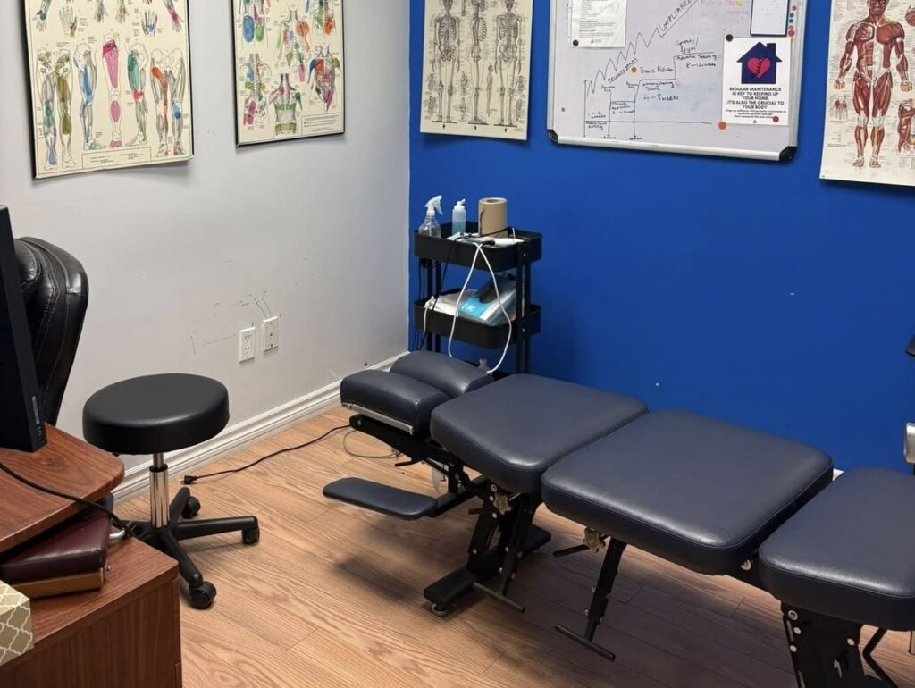 Modern physiotherapy clinic treatment room with blue walls, anatomical charts, framed credentials, and black padded treatment table.